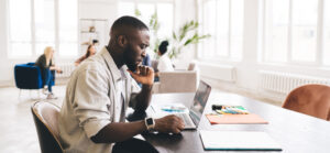 Side view of African American male employee in casual clothes sitting at table while touching chin and looking at laptop screen while thinking about project
