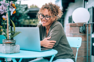 Happy woman using computer for video call outside home in the garden sitting on a table. Alternative office workplace outdoor.